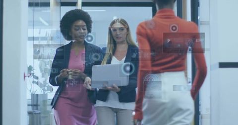 Diverse businesswomen walking through glass corridor holding laptop with futuristic UI overlays