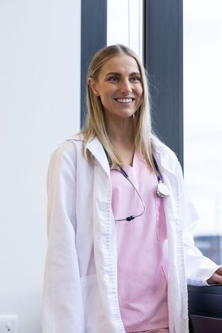 Smiling female doctor in white coat and pink scrubs with stethoscope by clinic window