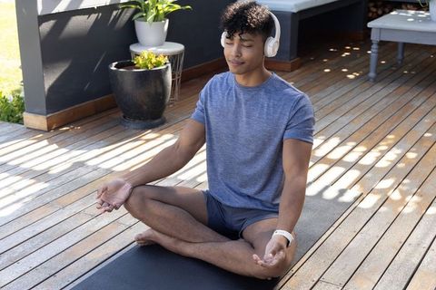 Young man meditating on patio deck wearing headphones and smartwatch in dappled sunlight