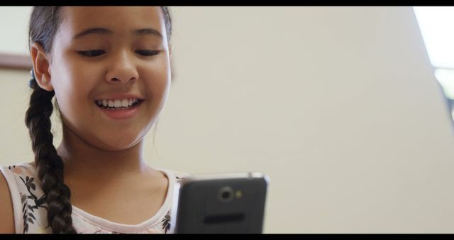 Young girl with long braided hair smiling while using a smartphone indoors. Ideal for images related to childhood, technology use among kids, digital communication, e-learning, and children's entertainment.