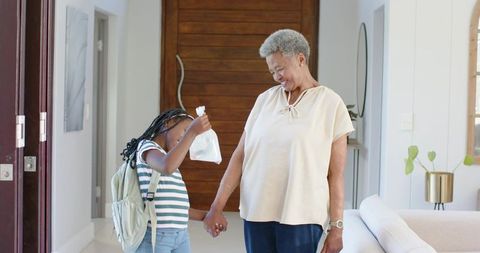 Grandmother holding hands with granddaughter in foyer, girl raising snack bag by door