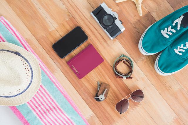 Travel essentials including a passport, smartphone, camera, sunglasses, hat, shoes, keys, bracelet, and towel laid out on a wooden board. Ideal for travel blogs, vacation planning websites, and summer adventure promotions.