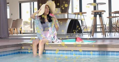 Young woman sitting poolside wearing straw sunhat and pastel kimono holding watermelon