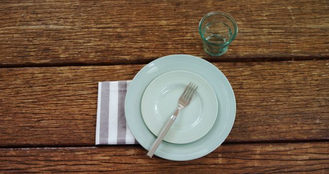 A simple table setting with an empty plate, fork, and a glass on a wooden surface, with copy space. It evokes a sense of anticipation for a meal or represents the concept of hunger and waiting.