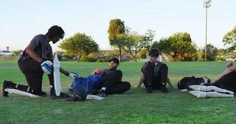 Cricket team preparing for practice on park field with african american man strapping pads