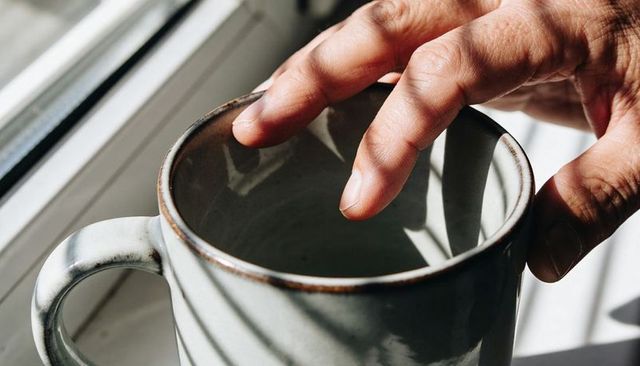 Adult hand touching ceramic mug rim on sunlit windowsill casting diagonal bold shadows