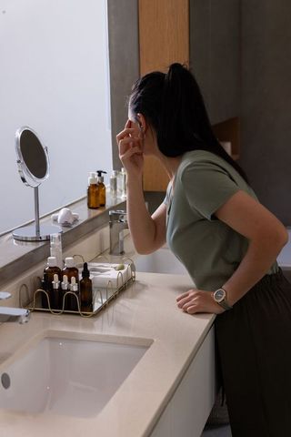 Mature african american woman leaning over bathroom vanity touching face amber skincare bottles