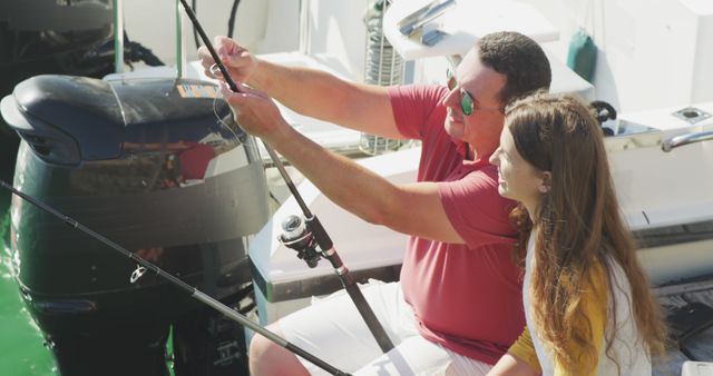 Side view of a Caucasian man and his teenage daughter enjoying time together on holiday in the sun by the coast, sitting on a jetty, holding fishing rods, the father is explaining how to use them