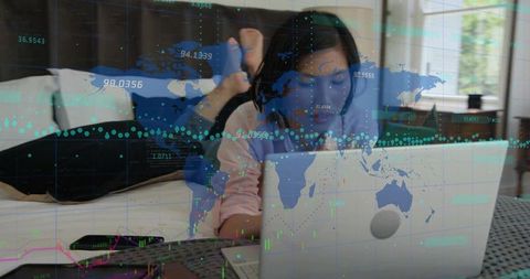 Reclining korean woman working on laptop on bed with world map and financial data overlay