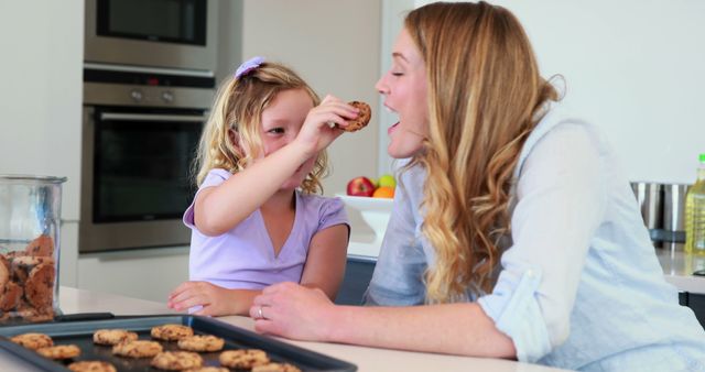 Little girl and her mother share a delightful moment together at home, enjoying cookies while seated in a cozy kitchen. This stock photo is ideal for family-related content, articles about parenting, lifestyle blogs, kitchen décor websites, and advertisement for home snacks or family-friendly products.