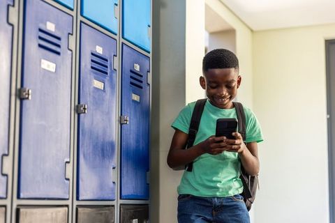 African american schoolboy leaning on lockers using smartphone smiling in hallway