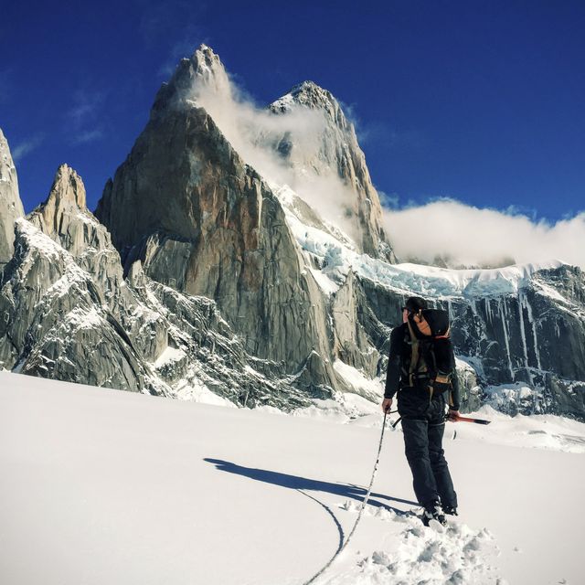 Mountaineer is walking across snow-covered mountain with ice axe in hand, towering peaks and clear blue sky in the background. Ideal for advertising adventure travel, outdoor gear, climbing equipment, or extreme sports. Can be used in articles about mountaineering, personal journey stories, or inspirational content about challenging endeavors.
