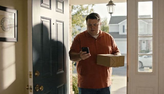 Checking smartphone while holding delivery package at suburban front door in daylight