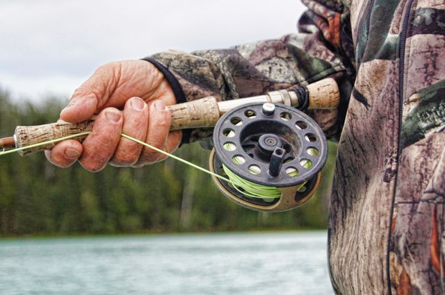 Close-up of a person dressed in camouflage jacket holding a fishing rod by a river. Useful for depicting outdoor activities, sports, hobbies, and nature-related content. Suitable for articles or promotions related to fishing equipment, outdoor adventures, or leisure activities.