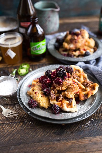 Waffles topped with a mixture of blackberries, raspberries, and deep-fried fritters, with syrup drizzled and powdered sugar. Picture of a wooden table in a rustic setting, cups, and a bottle in the background, suggesting a cozy morning breakfast. Perfect for food blogs, breakfast menus, recipe books, and advertisements for eateries.