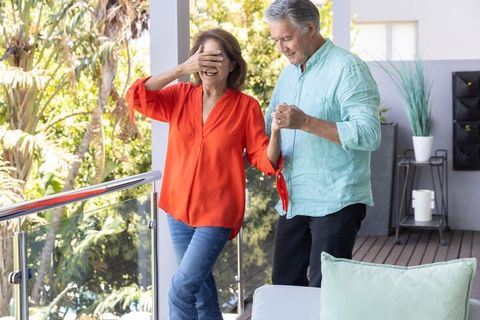 Mature couple sharing blindfold surprise on sunny balcony surrounded by tropical greenery