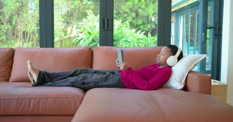 African american boy relaxing on tan leather sofa listening with headphones and tablet