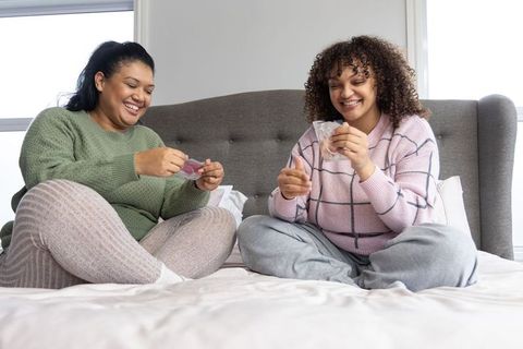Diverse female friends sitting on bed unwrapping wellness packets and laughing together