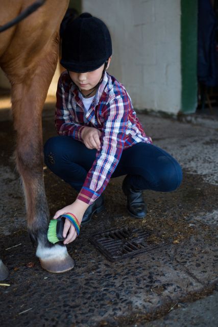Young girl wearing a helmet and plaid shirt, attentively brushing a horse's leg in a stable. Ideal for use in content about animal care, equestrian activities, children learning responsibilities, farm life, and horsemanship training. Perfect for websites, magazines, or educational materials focusing on parenting, farming, or horse riding.