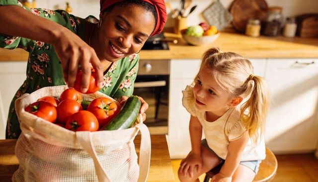 Parent and child unpacking fresh tomatoes and cucumber from reusable mesh bag in sunlit kitchen