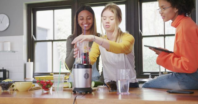 Group of friends blending a healthy smoothie together in a modern kitchen. They are gathered around a blender, preparing fruit and enjoying the activity. Ideal for content related to healthy living, cooking, friendship, lifestyle blogs, and kitchen appliances promotions.