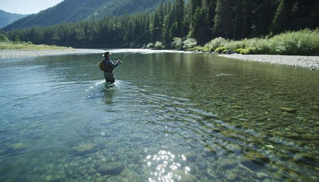 Fly fisher casting in crystal-clear mountain river wearing waders and brimmed hat