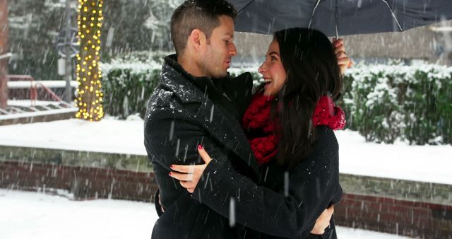 Couple standing close under umbrella with snow falling, both dressed warmly in dark outerwear and sharing an affectionate moment. Ideal for depicting winter romance, love stories during the holiday season, or being used in advertisements for winter clothing, romantic getaways, or holiday greeting cards.