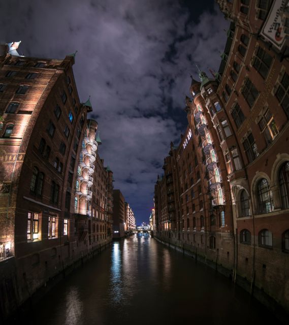 Nighttime perspective of a historic European canal district with illuminated brick buildings on both sides. Cloudy sky reflects in the calm waters below, creating a scenic and tranquil atmosphere. Ideal for use in travel blogs, tourism promotions, architectural studies, and historical presentations.