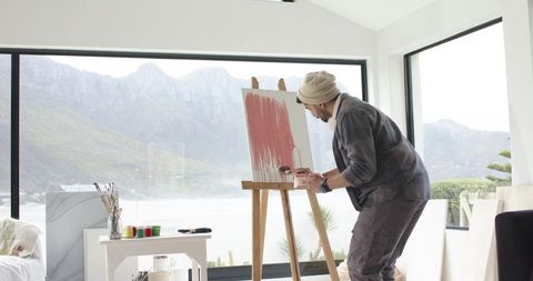 Indian man painting on easel in bright mountain-view studio with windows, coastal view