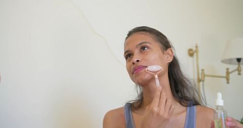 Young woman massaging jaw with rose quartz facial roller while holding serum dropper