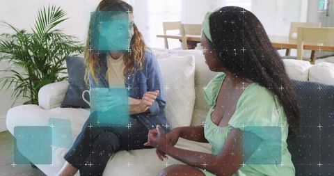 Two women chatting on couch holding mug with teal digital overlays cozy living room scene
