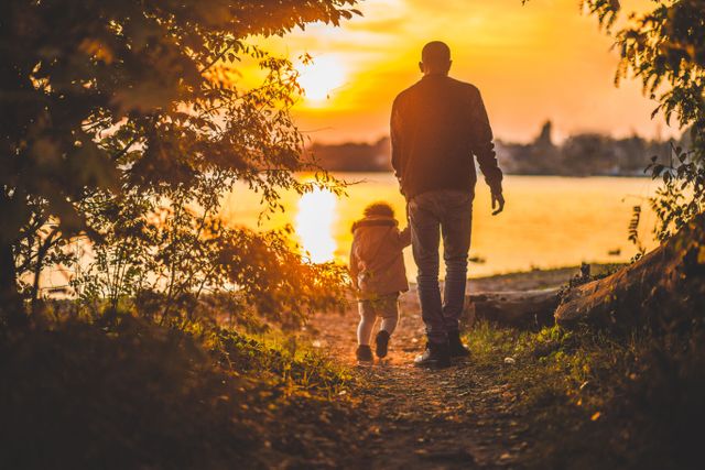 Silhouette of father and child walking hand in hand along a lakeside at sunset. Perfect for uses in parenting blogs, family outdoor activity promotions, bond-building campaigns, or inspirational content that emphasizes family connections and the beauty of nature.