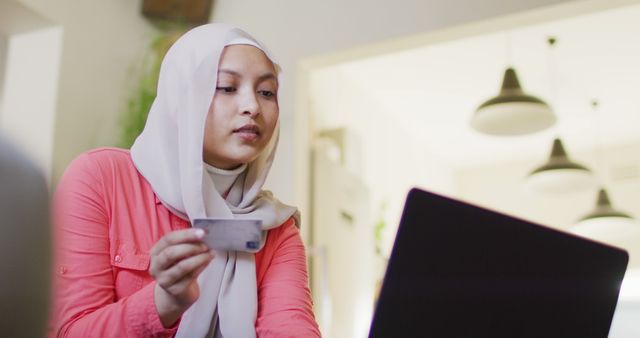 Image of smiling biracial woman in hijab making online payment using laptop and credit card at home. Happiness, communication, finance, inclusivity and domestic life.