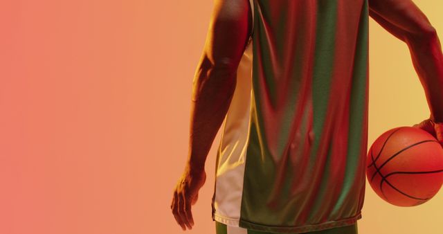 Athletic man holding basketball wearing green and white jersey against vibrant gradient background. Ideal for use in sports advertisements, fitness promotions, and basketball-related content.