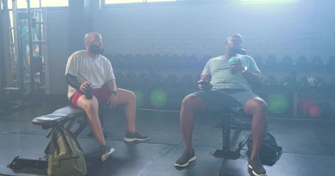 African American Workout Partners Sitting and Chatting Between Sets by Dumbbell Rack in Sunlit Gym