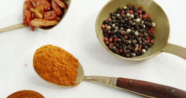 Close-up of various spices and seasonings in metallic spoons on a white background. Includes a spoonful of mixed peppercorns, turmeric powder, and chili flakes. Ideal for use in culinary blogs, cooking websites, recipe books, and food-related advertising to showcase the richness and diversity of ingredients.