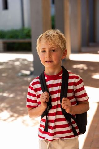 Blond schoolboy wearing red white striped t-shirt carrying backpack in sunlit walkway