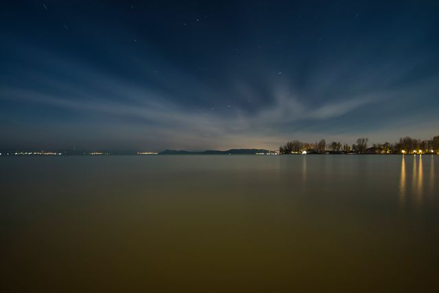 Serene night view showing calm lake reflecting the vast sky above with distant urban lights illuminating the horizon. Perfect for use in backgrounds, nature and travel blogs, or relaxation and meditation apps emphasizing tranquility and peace.