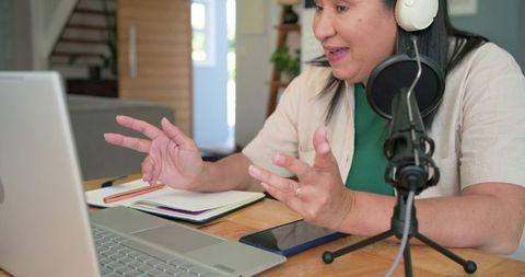 Mature woman recording podcast at home desk with laptop and condenser microphone