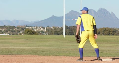 Baseball player standing on base in yellow and blue uniform holding glove with mountain backdrop