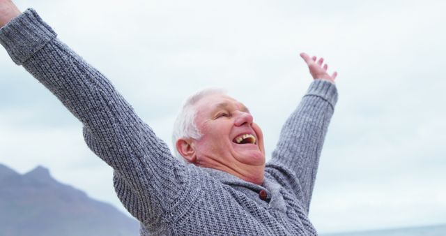 Senior man wearing grey sweater smiling joyfully with arms raised outdoors. Background includes mountains and sky, suggesting nature and freedom. Perfect for themes such as active aging, happiness, outdoor activities, well-being, and joy.