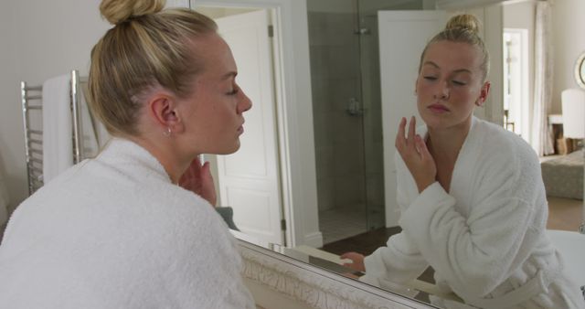 Woman is standing in front of a bathroom mirror and applying skincare products, dressed in a white bathrobe. Emphasizes themes of self-care, beauty, and morning routines. Useful for beauty blogs, personal care product promotions, and articles focusing on everyday grooming habits.