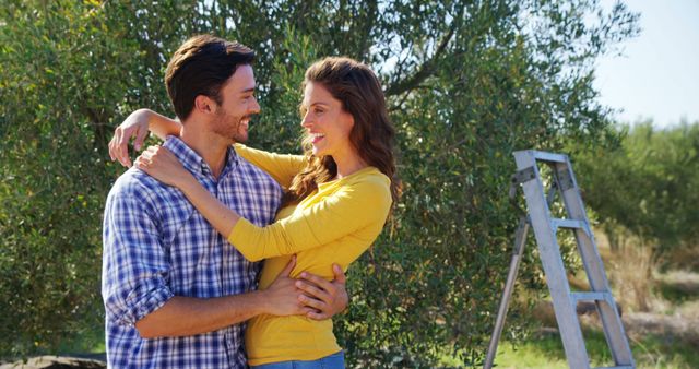 A young Caucasian couple embraces lovingly in an orchard, with a ladder in the background suggesting a day of fruit picking. Their affectionate pose and joyful expressions convey a sense of romance and companionship amidst a serene natural setting.