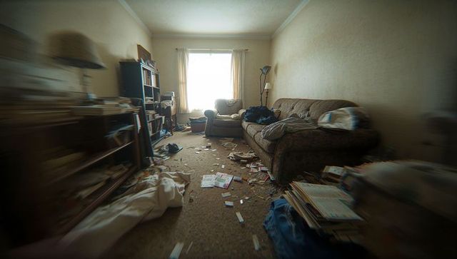 Cluttered living room with scattered papers, worn sofa and armchair under sunlit window