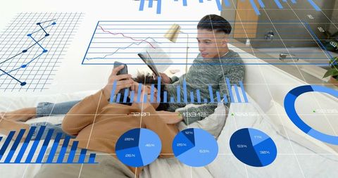Two men reclining on bed sharing tablet and smartphone viewing financial charts overlay