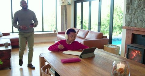 African american father guiding son remote learning on tablet at home dining table