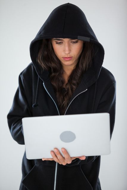 Young adult wearing a black hoodie, concentrating while using a laptop. Ideal for illustrating concepts related to cybersecurity, hacking, technology, and internet security. Useful for articles, blogs, and presentations on online safety, coding, and digital threats.