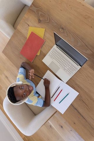 African american boy studying at home wearing headphones and using laptop while writing in notebook
