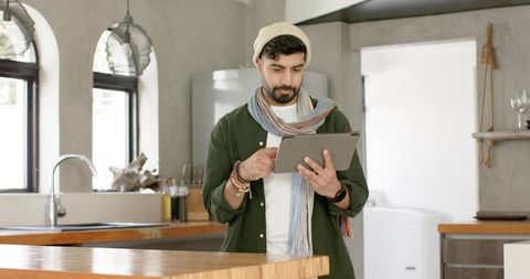 Indian man using tablet in minimalist modern kitchen, working remotely and browsing ideas