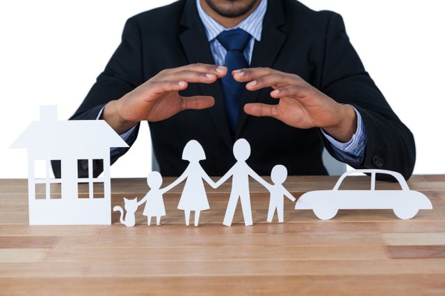 Businessman in suit and tie protecting paper cut out figures of a family, house, and car with hands. Ideal for illustrating concepts of insurance, financial protection, safety, and security. Useful for insurance company advertisements, financial services promotions, and articles on family protection.